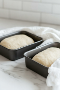This image shows the risen dough being shaped into loaves and placed in loaf pans, preparing for the final rise before baking.