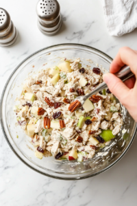This image shows salt and black pepper being sprinkled over the creamy chicken salad mixture, enhancing the flavors and adding a perfectly balanced seasoning to complement the sweet and savory ingredients.