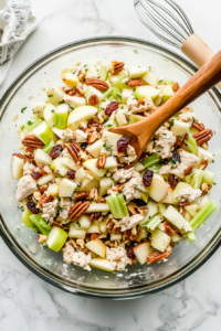 This image shows a spoon stirring the chicken salad mixture, ensuring that the creamy dressing evenly coats the canned chicken, apples, pecans, celery, and raisins for a well-blended and flavorful result.