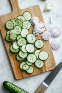 This image shows freshly harvested cucumbers and onions being sliced into thin rounds on a cutting board, preparing them for the sweet pickling process.