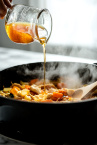 This image shows a stream of rich, golden chicken broth being poured into a pot, blending with seared vegetables to create a flavorful and nourishing base.