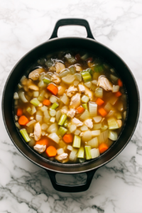 This image shows a whole chicken simmering in a large pot of golden broth alongside carrots and celery, releasing deep, savory flavors into the soup base.