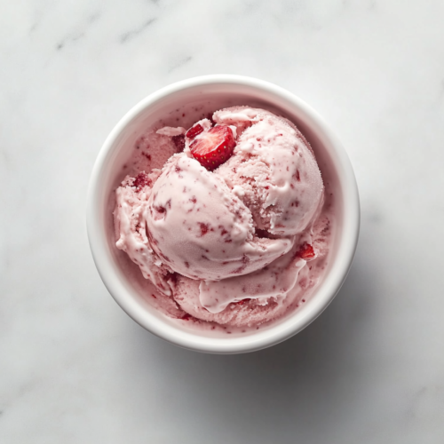This image shows a scoop of creamy pink Strawberry Ice Cream served in a round white ceramic bowl, garnished with small pieces of fresh strawberries for a delightful presentation.