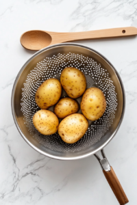 This image shows boiled potatoes being drained in a colander, allowing them to cool slightly before peeling and chopping.