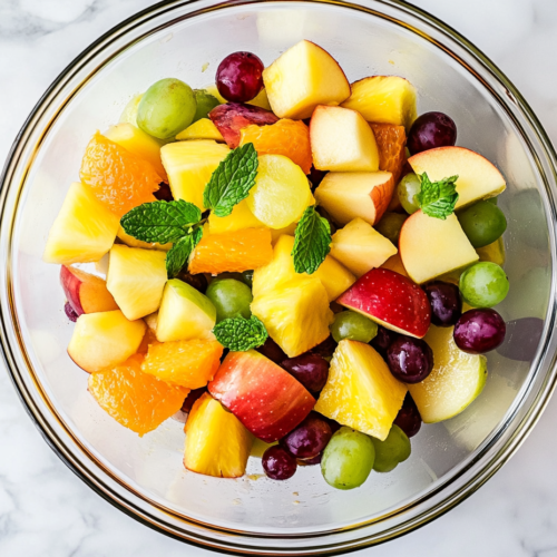 This image shows a vibrant fruit cocktail salad in a glass bowl, featuring a colorful mix of chopped pineapple, mandarin oranges, grapes, apples, and assorted berries. Fresh basil leaves are sprinkled on top, enhancing the flavor and presentation.