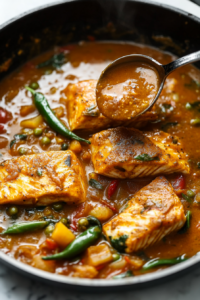This image shows fresh fish pieces being carefully placed into the simmering curry, absorbing the rich flavors of the spices and tamarind-soaked liquid. The fish remains intact while gently cooking in the aromatic broth.