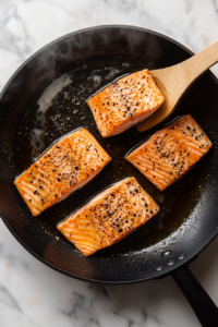 This image shows a salmon fillet cooking in a hot pan, sizzling as it develops a golden-brown crust while staying tender inside.