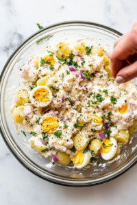 This image shows salt and freshly ground black pepper being sprinkled over the potato salad to enhance its taste with a final seasoning touch.