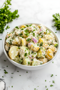 This image shows a serving of creamy potato salad in a white bowl, garnished with fresh herbs and ready to be enjoyed.