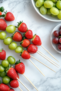 This image shows strawberries and grapes being carefully threaded onto wooden skewers. The fruit is evenly spaced to allow for an even coating of the sugar syrup.