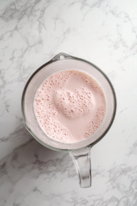 This image shows a top-down view of a blender filled with a smooth pink strawberry ice cream mixture, blending the ingredients into a creamy base