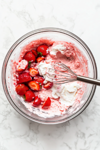 This image shows a top-down view of a mixing bowl filled with fresh strawberries, cream, sugar, and other ingredients, ready to be combined for homemade strawberry ice cream.