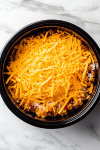 This image shows a generous layer of shredded cheese being sprinkled over the top of the casserole mixture, ready to melt into a gooey, cheesy topping.