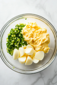 This image shows chopped eggs combined with creamy mayonnaise, tangy mustard, and sliced green onions in a white mixing bowl, ready for seasoning.