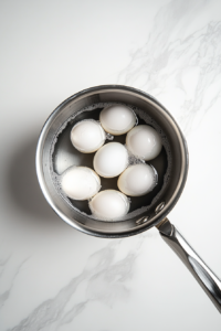 This image shows whole raw eggs resting in a saucepan filled with cold water, ready to be brought to a boil for egg salad preparation.