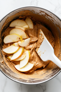 Thin apple slices being gently folded into the cake batter with a spatula.