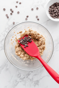 This image shows a top-down view of a mixing bowl where semi-sweet chocolate chips are being gently folded into the peanut butter oat mixture with a spatula, ensuring an even distribution of chocolatey goodness in each bite.