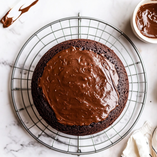 This image shows a freshly baked, fluffy chocolate cake with smooth ganache frosting, decorated with chocolate shavings, placed on a round cake stand over a clean white marble countertop with a simple background.