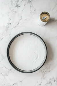 This image shows the process of lightly greasing a round cake pan with non-stick spray on a white marble countertop.