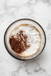 This image shows a glass mixing bowl on a white marble countertop with flour, sugar, cocoa powder, baking powder, and salt for the chocolate cake.