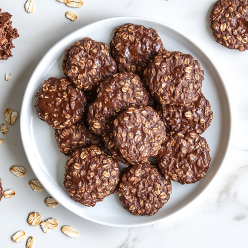 This image shows a plate of round, no-bake chocolate oatmeal cookies with visible oats infused throughout, creating a rustic and textured appearance. The cookies are arranged on a simple white plate, highlighting their rich chocolatey color and homemade charm.