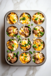 This image shows chopped green onions, shredded cheddar and Swiss cheese, and cooked sausage being sprinkled evenly into the greased mini muffin cups, creating colorful and tasty layers.