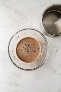 This image shows a top-down view of thin and fluid chocolate cake batter in a clear glass mixing bowl after boiling water has been stirred in, placed on a white marble countertop.