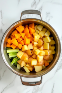 This image shows chopped onion, celery, carrot, potatoes, and butternut squash in a stainless steel pot with melted butter, all placed on a white marble cooktop.