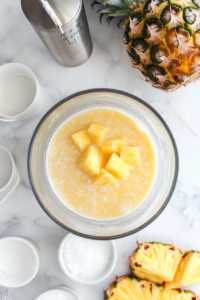 A blender filled with coconut milk, pineapple juice, white rum, and sugar, ready to be blended into a tropical drink.