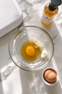 This image shows an egg being added to a creamy butter-sugar mixture inside a clear glass bowl, with a bottle of vanilla extract and a white electric hand mixer resting nearby on a white marble cooktop.