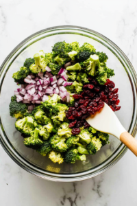 This image shows chopped red onions and dried cranberries being added to the broccoli, bringing color and tangy sweetness to the mix.