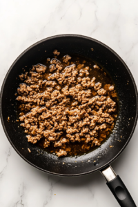 adding-seasonings-and-liquids-to-browned-chicken-in-black-skillet-on-white-marble