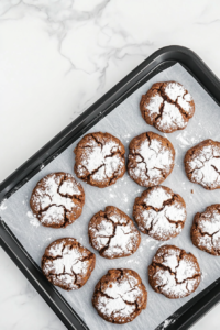 This image shows freshly baked chocolate crinkle cookies on a parchment-lined black baking sheet resting on a white marble countertop.