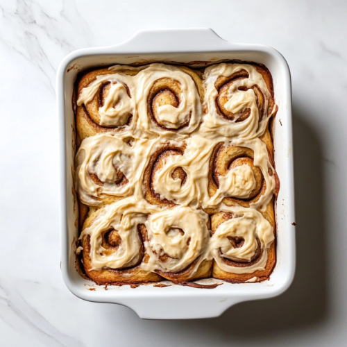 This image shows a white baking dish filled with golden-brown banana bread cinnamon rolls, topped with a thick layer of creamy frosting.