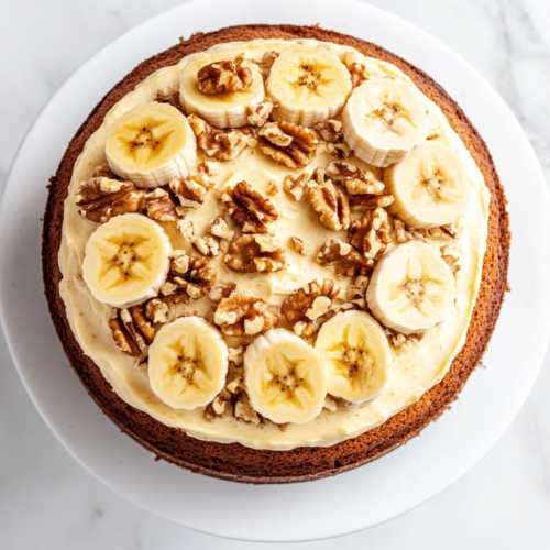This image shows a perfectly round Banana Walnut Cream Cake, golden brown and crowned with fresh banana slices and chopped walnuts, creating a rustic yet elegant dessert centerpiece.