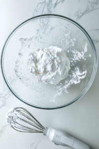 This image shows a large glass bowl with stiffly beaten egg whites, cream of tartar, and sugar on a white marble countertop with a white electric hand mixer.