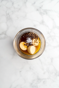 This image shows a glass blender filled with balsamic vinegar, olive oil, honey, Dijon mustard, and diced shallots being blended into a smooth, glossy dressing.