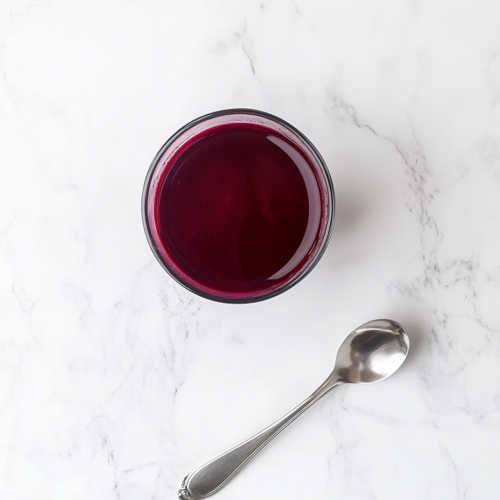 This image shows a striking glass of deep red beetroot juice placed on a light surface with a silver spoon resting beside it, highlighting the juice’s rich color and smooth texture, ready to be enjoyed.