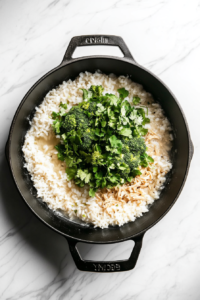 This image shows bright green broccoli florets added on top of simmering rice and broth in a black skillet on a white marble countertop.