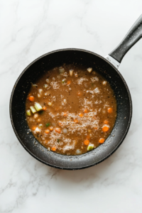 This image shows a black skillet on a white marble cooktop, with chicken broth added to the vegetable mixture, bubbling and boiling.