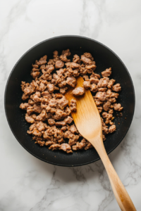 This image shows sausage browning in a skillet, being broken into crumbles with a spatula, releasing its rich, savory aroma.