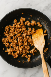 This image shows ground chicken being browned in olive oil in a black nonstick skillet placed on a white marble cooktop, with a wooden spatula resting on the side.