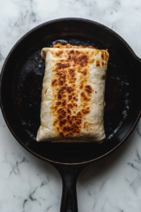 This image shows a rolled burrito placed seam-side down on a hot black cast iron skillet over a white marble cooktop, ready for grilling.