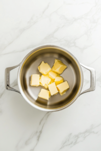 This image shows a stainless steel pot placed on a white marble cooktop with cubes of unsalted butter beginning to melt over medium heat.