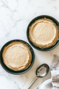 This image shows two black round cake pans filled with cake batter on a white marble cooktop, with a silver ice cream scoop placed nearby.