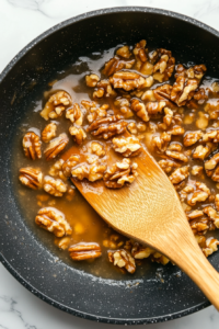 This image shows chopped walnuts sizzling in a buttery brown sugar mixture in a skillet, beginning to caramelize and take on a glossy golden coat.