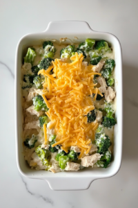 This image shows a white ceramic casserole dish filled with creamy chicken and rice mixture and topped with shredded cheddar cheese, placed on a white marble countertop.