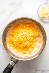 This image shows a pat of cheese melting in a shiny stainless steel saucepan placed on a clean white marble cooktop, with a small clear glass bowl of butter beside it.
