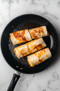 This image shows cheesy chicken wraps placed seam-side down in a black non-stick skillet, toasting on a white marble cooktop.