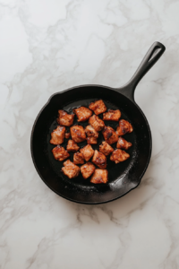 This image shows seasoned chicken breasts sizzling on a black cast iron skillet over a white marble cooktop. The chicken is cooking and slightly charred.